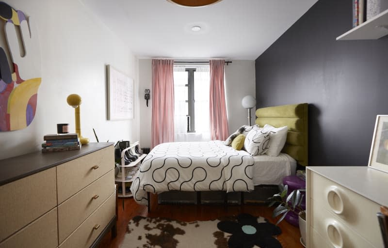 A bedroom with one black feature wall, pink curtains, and a neutral dresser. A green fabric headboard is behind a queen bed.