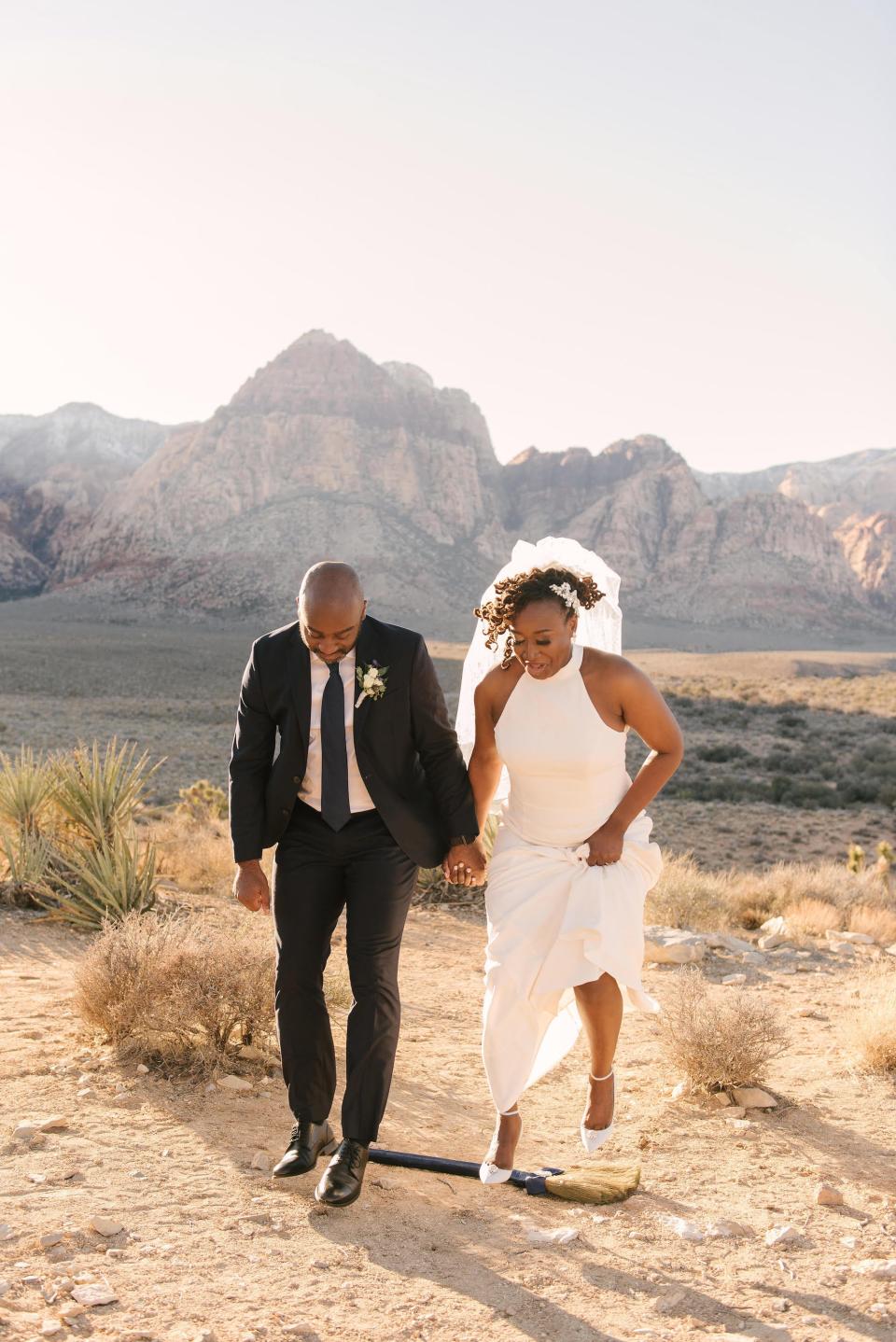 A bride and groom jump the broom in a desert.