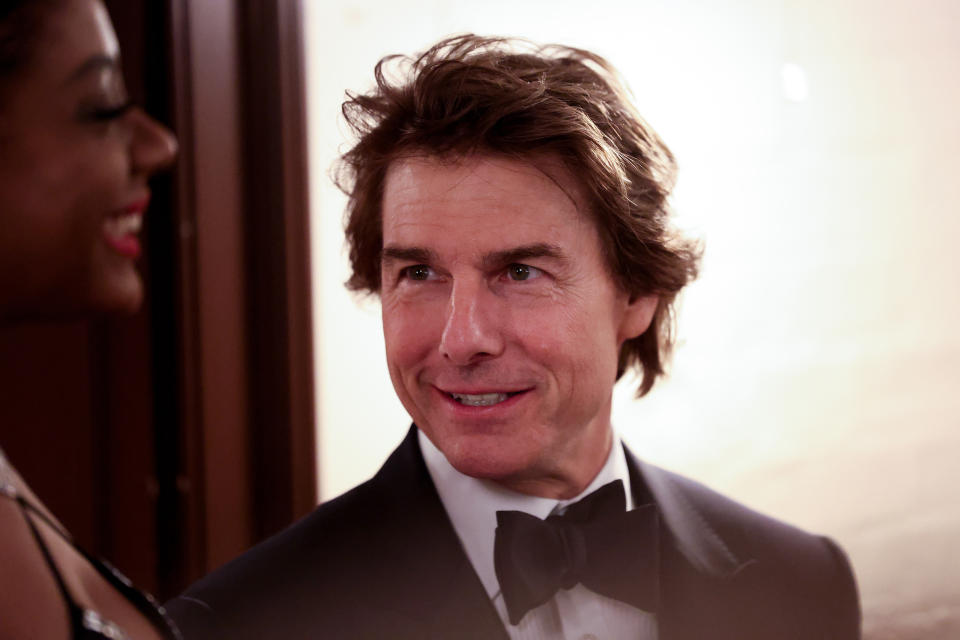 Tom Cruise, dressed in a formal black suit and bow tie, smiles at a woman out of focus in the foreground