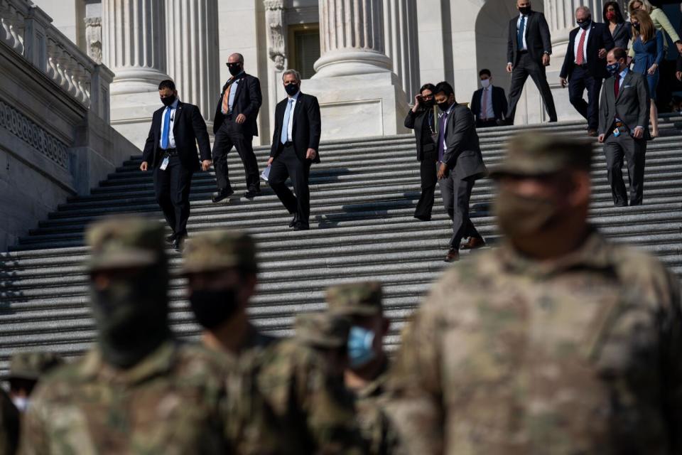 Several people in suits walk down stairs outside a building while people in military garb are in the foreground.