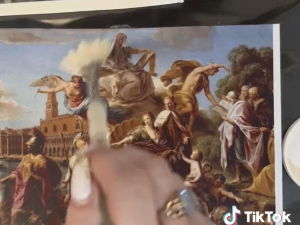 screenshot of a hand holding a paint brush over a print-out of a painting.