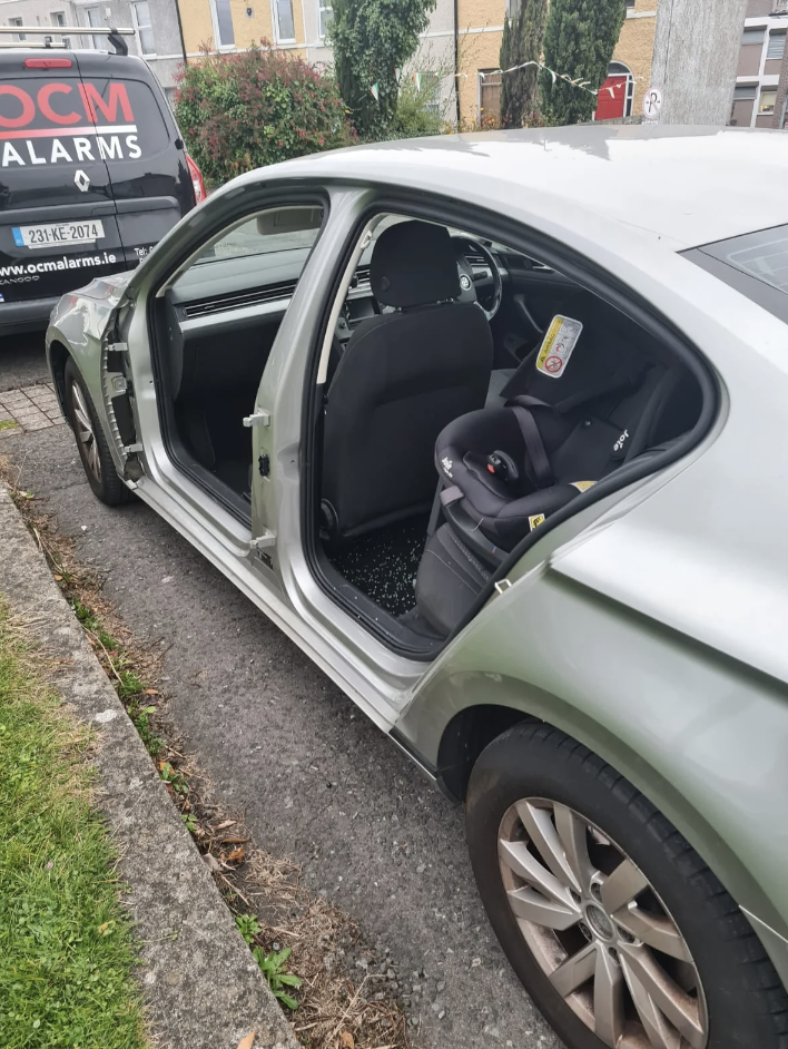 Car door open, revealing a child's car seat inside. Vehicle parked by roadside with another car's reflection visible