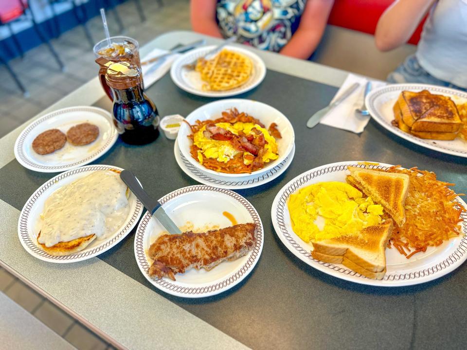 A gray table filled with white plates containing food. The plates hold biscuits and gravy, a piece of steak, eggs, toast, and hash browns, a hash-brown bowl, and a waffle.