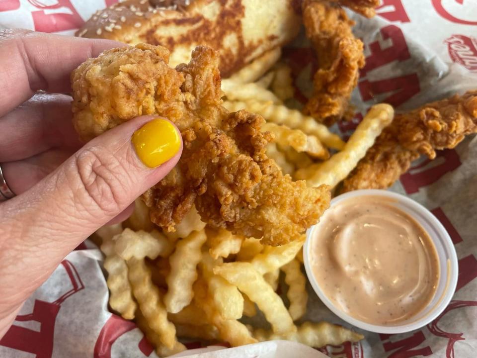 The writer holds a chicken finger in front of sauce and fries at Raising Cane's