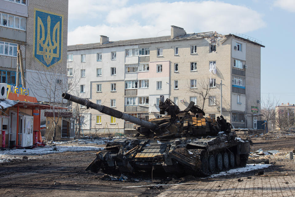A burned tank in front of a building bearing a yellow-and-gold Ukrainian insignia, with a bombed apartment building behind.