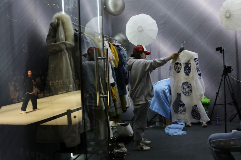 Salesperson promotes clothings by XUNRUO during a live streaming session, inside a booth while a model onstage is presenting creations from CHUSAN designed by Guoqin Zhang, during China Fashion Week, in Beijing
