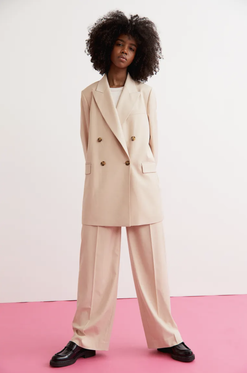 black model with afro standing on pink floor wearing beige wide leg pants and beige Double-Breasted Jacket (Photo via H&M)