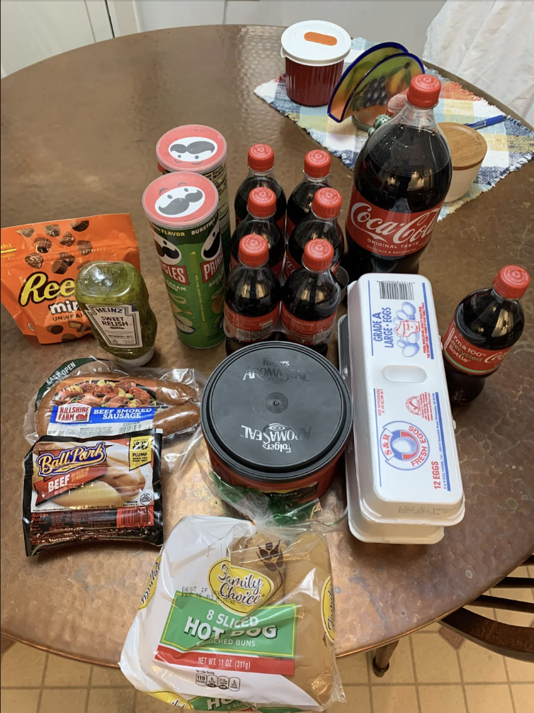 Various groceries on a table, including soda bottles, hot dog buns, eggs, beef hot dogs, Pringles, Reese's snacks, guacamole, and pimento cheese