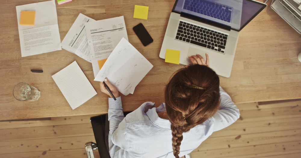 A new report has found that LGBTQ+ Women are discriminated in the workplace. The image is an overhead shot of a person with long plaited hair sitting at a table with papers and a laptop.