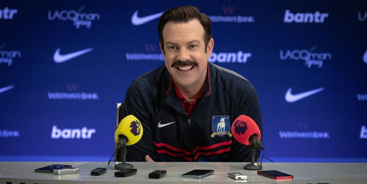 a publicity still of jason sudeikis portraying ted lasso, wearing a blue athletic jacket, sitting at a table with two microphones and several cellular phones and recording devices, with the words we are richmond in the front, and a blue backdrop with various advertising logos in the background