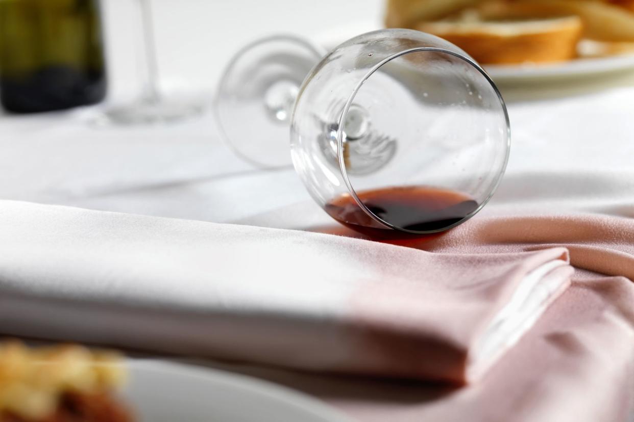 Focus on a toppled wine glass with red wine spilt on a white tablecloth next to a white napkin, both stained by red wine, surrounded by blurred food on plates on the table in both the foreground and background