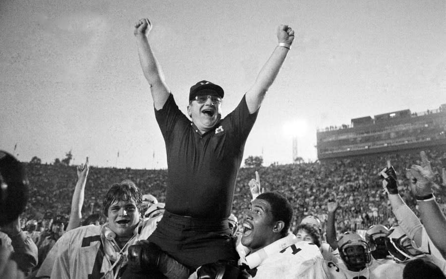 Bo Schembechler holds both of his arms in the air as he is carried off the field by two Michigan players.