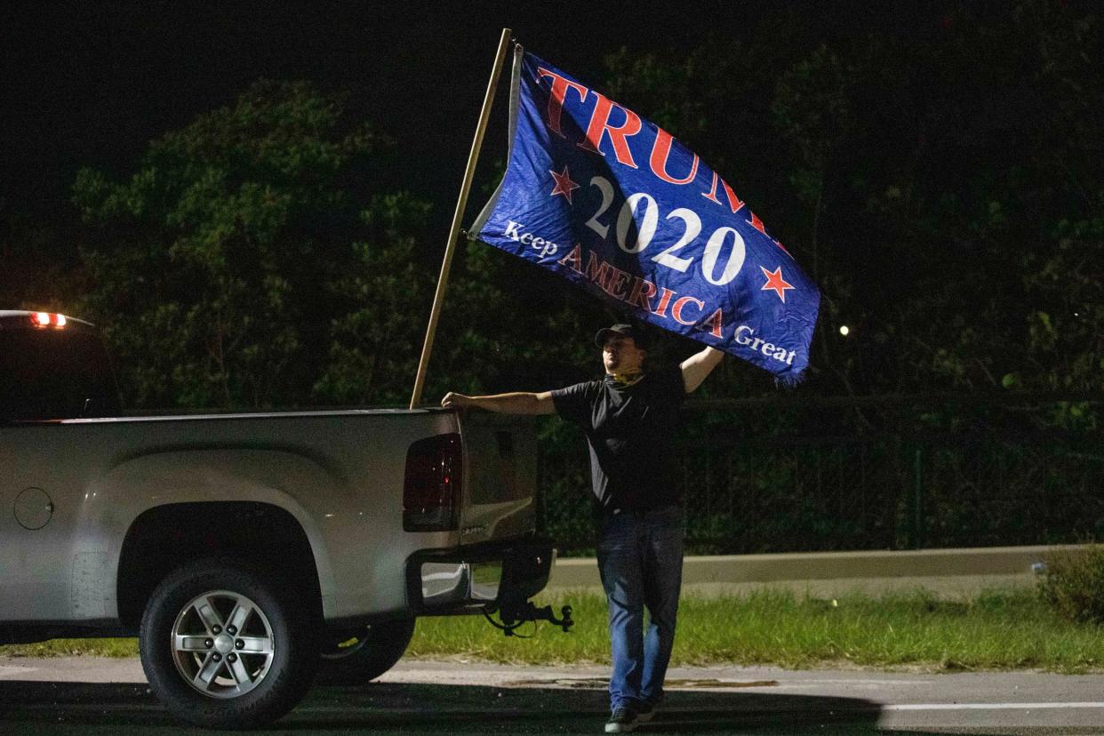 A man who declined to provide his name stands by a flag expressing support for former President Donald Trump near his Mar-a-Lago estate in Palm Beach, Fla., Monday, Aug. 8, 2022. 