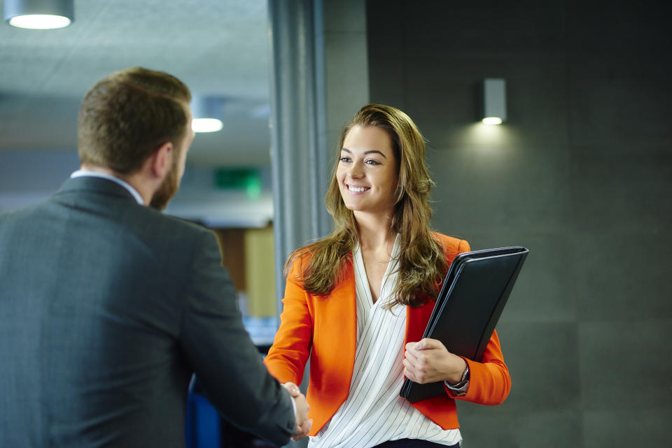 A young female job candidate is shaking hands with her prospective employer. She is smiling confidently and looking him in the eye . She is holding her cv resume . they are both standing in a modern office interior.