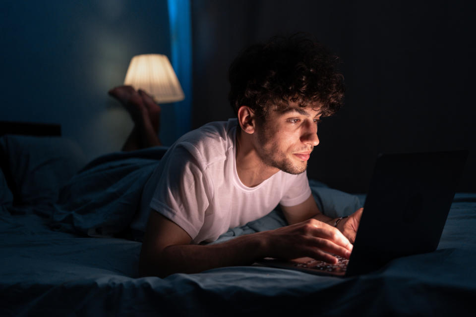 A person is lying in bed using a laptop at night. A lamp softly illuminates the scene