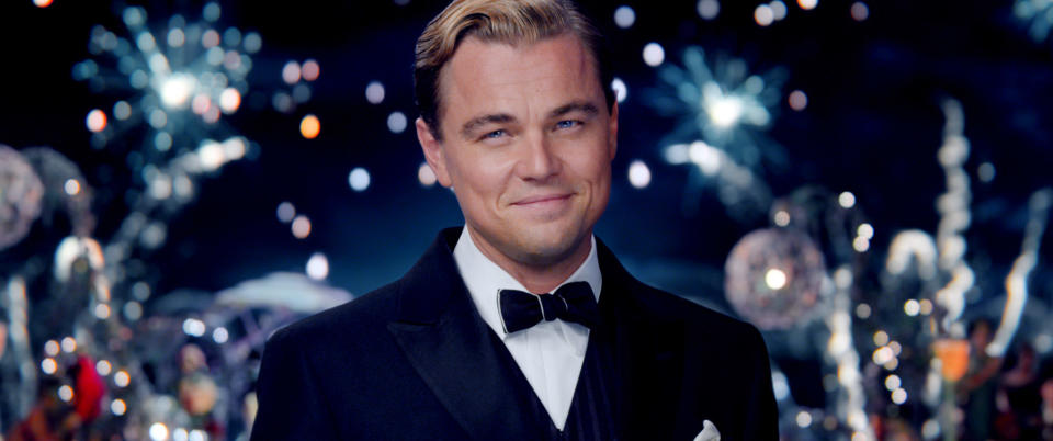 Leonardo DiCaprio in a tuxedo with a bow tie, smiling against a festive background with fireworks