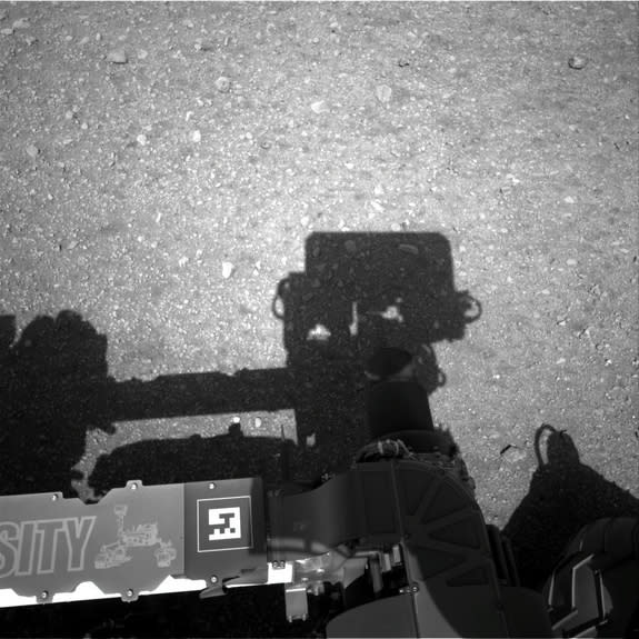 This is the first image taken by the Navigation cameras on NASA's Curiosity rover. It shows the shadow of the rover's now-upright mast in the center, and the arm's shadow at left. The arm itself can be seen in the foreground. Image released Aug