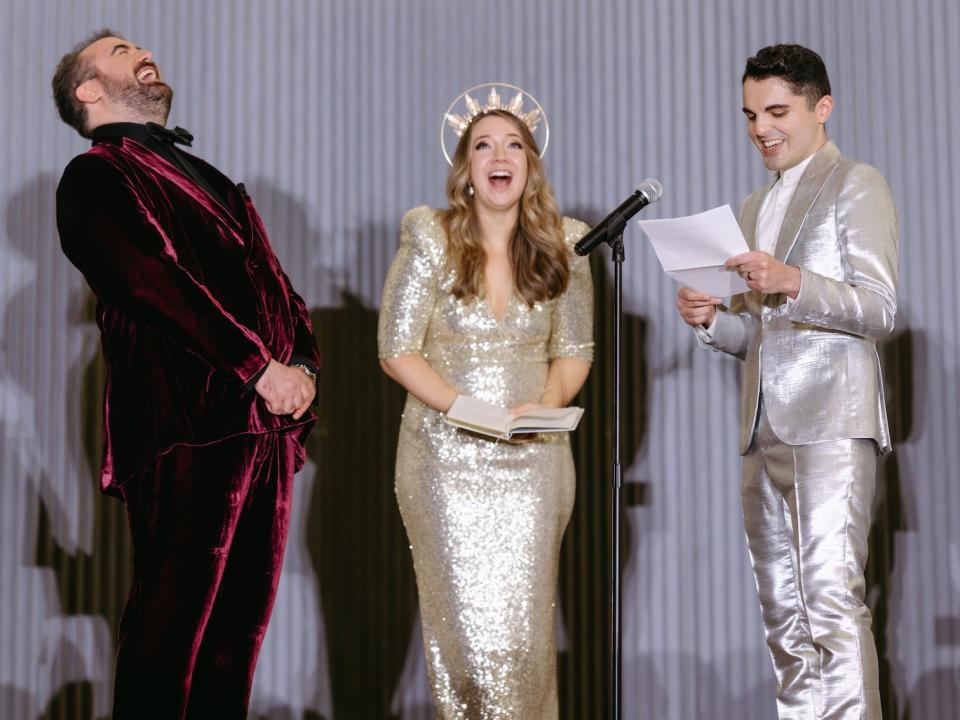 A groom laughs as his husband reads his vows. Their officiant stands between them in a gold dress wearing a crown.