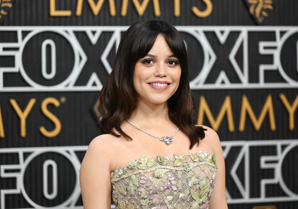 Jenna Ortega at the Emmys, wearing a strapless floral-patterned dress and a necklace, standing in front of a backdrop with "FOX" and "EMMYS" text