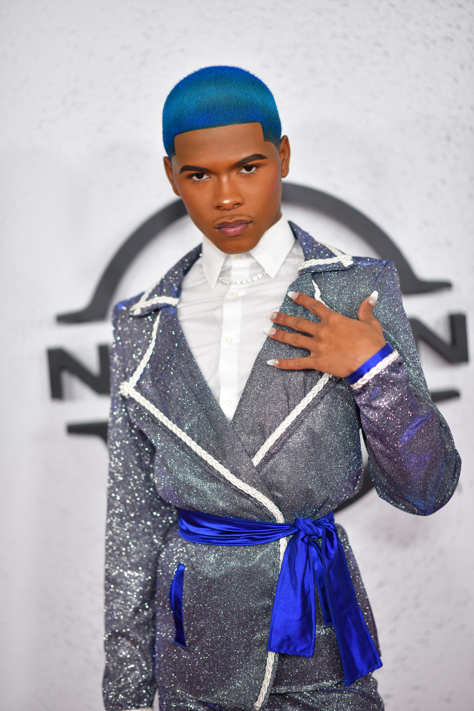 Kidd Kenn poses on carpet in a sparkly blazer with white shirt and a sash