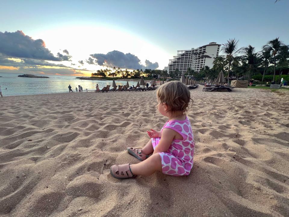 A baby girl sitting on a beach in a white and pink dress looking at the ocean.