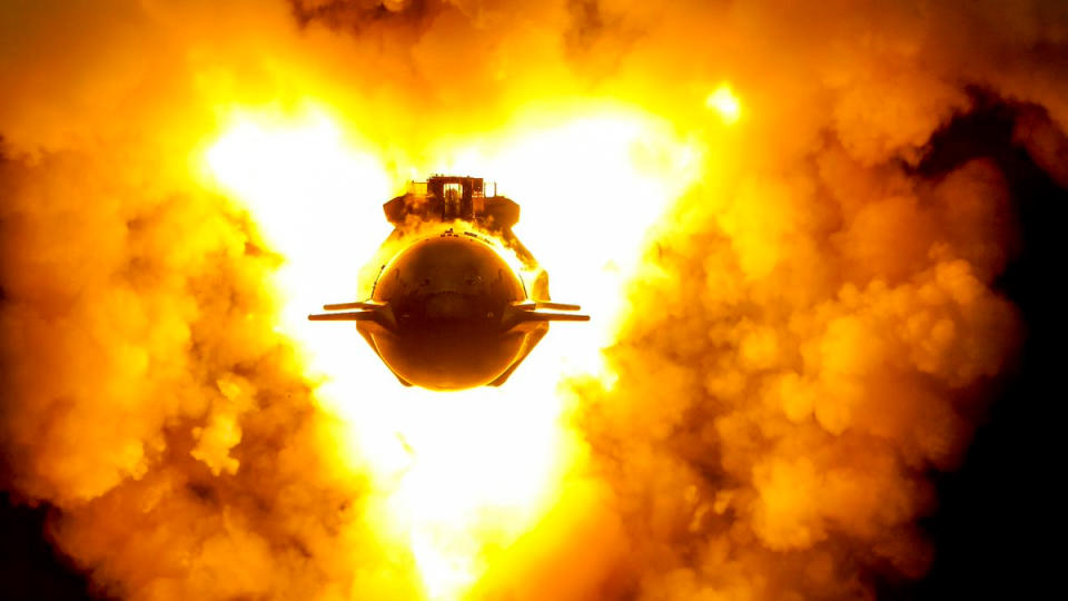 top-down view of shiny silver starship vehicle conducting an engine test, with orange flames and dust billowing around it