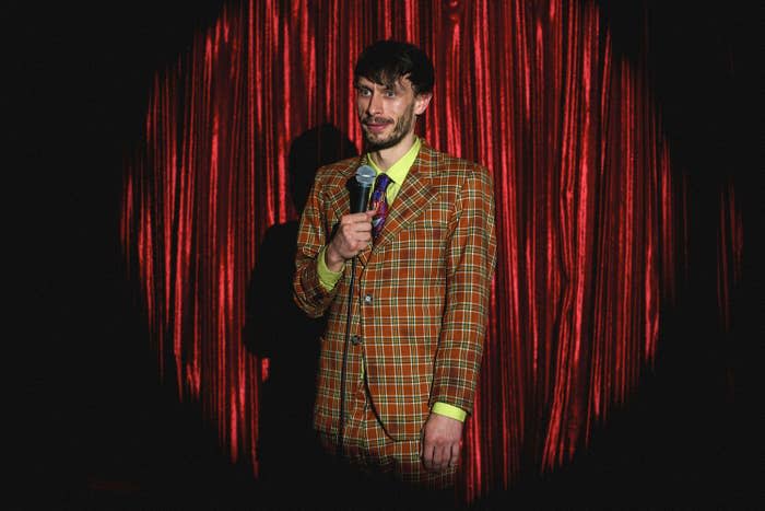 Richard Gadd in a plaid suit stands with microphone in front of red curtain