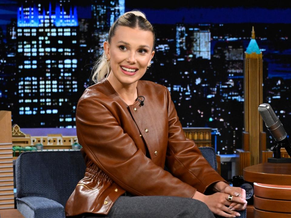 Millie Bobby Brown in a brown jacket and grey pants during a late-night interview with Jimmy Fallon