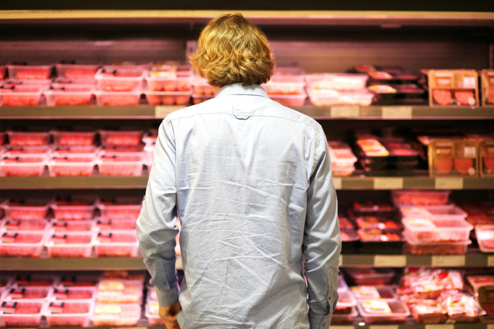 A person with shoulder-length blond hair wearing a light shirt stands in front of a grocery store meat section, looking at various packaged meats