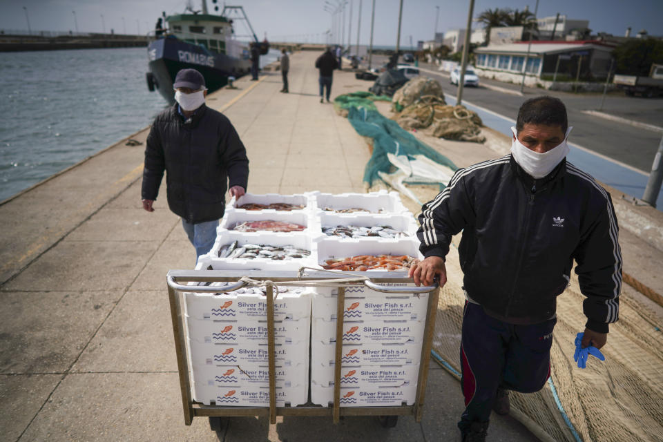 Workers, wearing protective masks, carry crates of fish to the Silver Fish wholesale auction house, at Fiumicino fishing port, on the outskirts of Rome Monday, March 30, 2020. Italy’s fishermen still go out to sea at night, but not as frequently in recent weeks since demand is down amid the country's devastating coronavirus outbreak. (AP Photo/Andrew Medichini)
