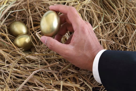 A business-suited arm reaches out to grab one of three golden eggs from a nest.