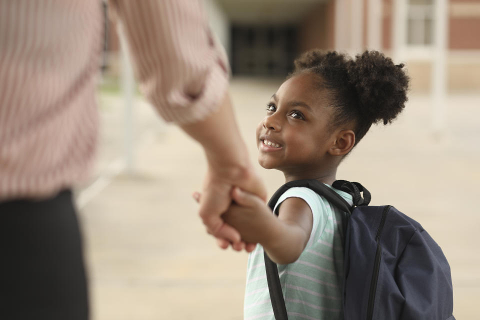 A child with a backpack holds an adult's hand, looking up and smiling as they walk together. The image conveys themes of guidance and support