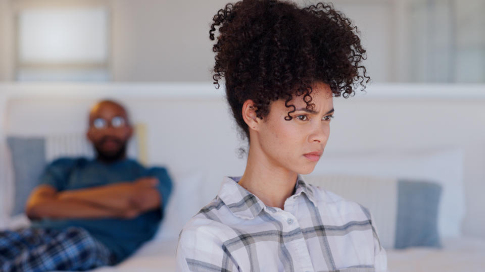 Woman looks upset in the foreground with a man sitting on a bed in the background, both indoors