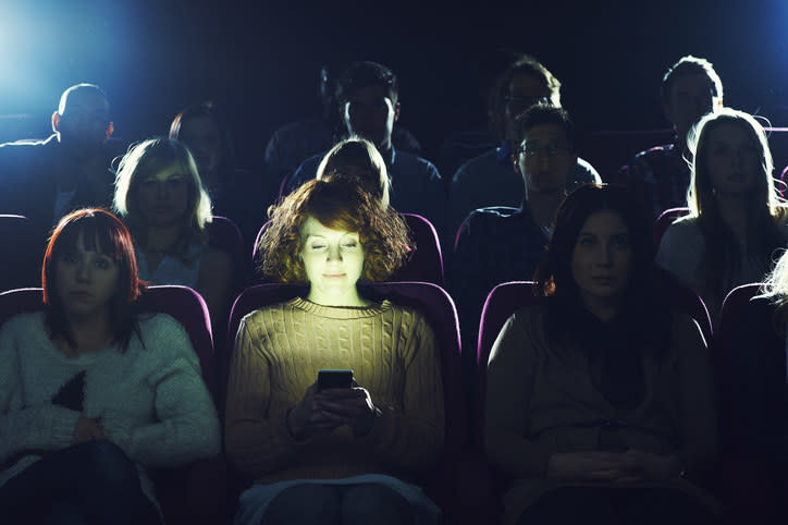 A person staring at their phone in a dark movie theater as other people who are trying to watch the film