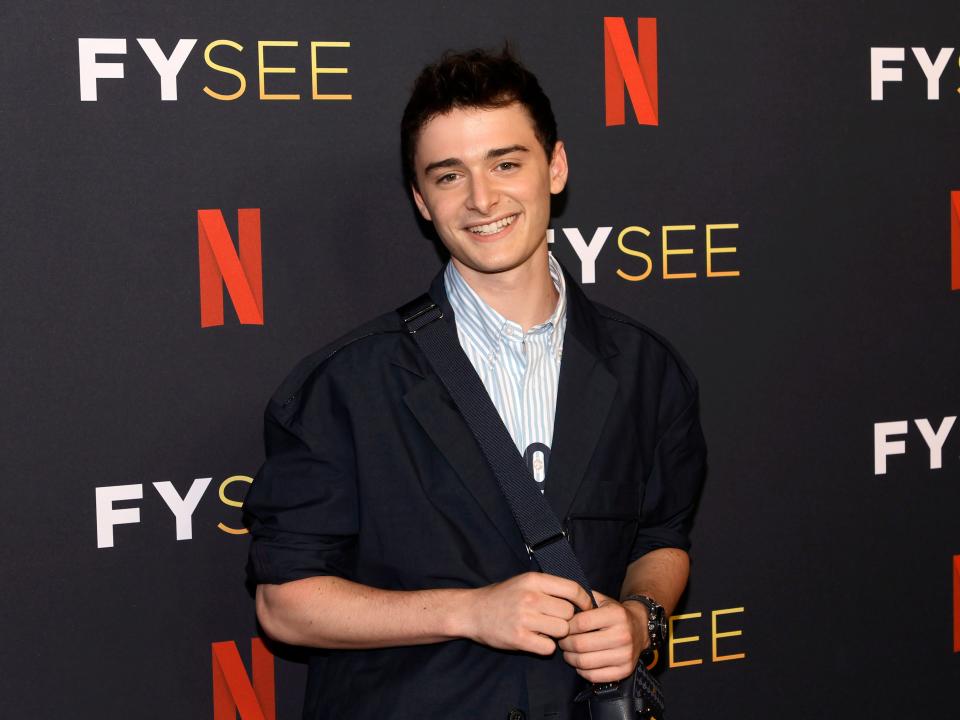 Noah Schnapp smiling and holding the strap of a satchel bag while standing in front of a Netflix-branded wall.