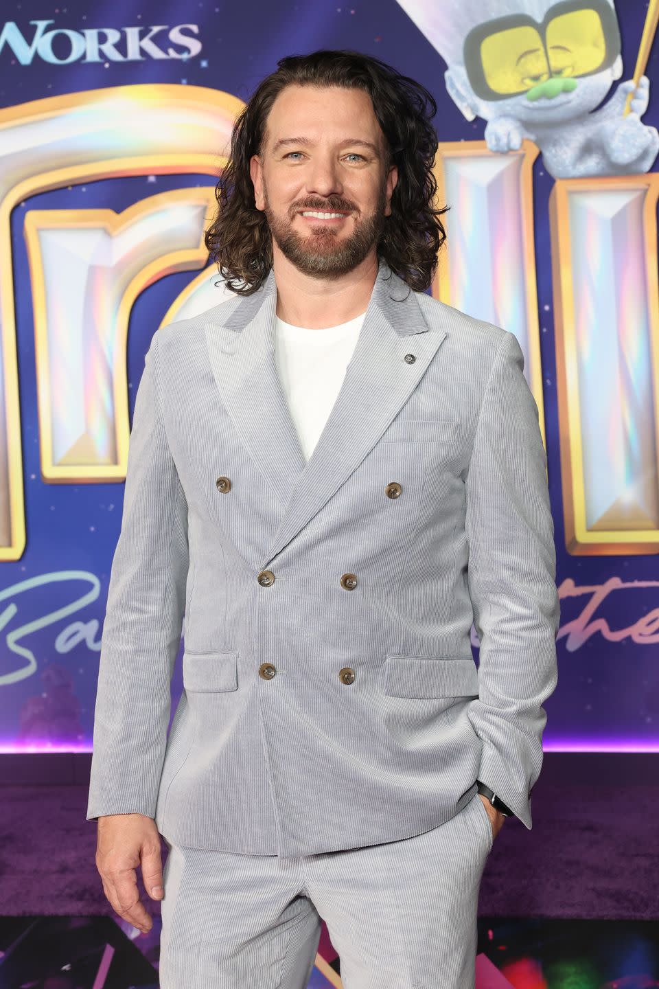 jc chasez wears a gray suit with a white shirt and smiles at the camera, he stands in front of a trolls movie poster