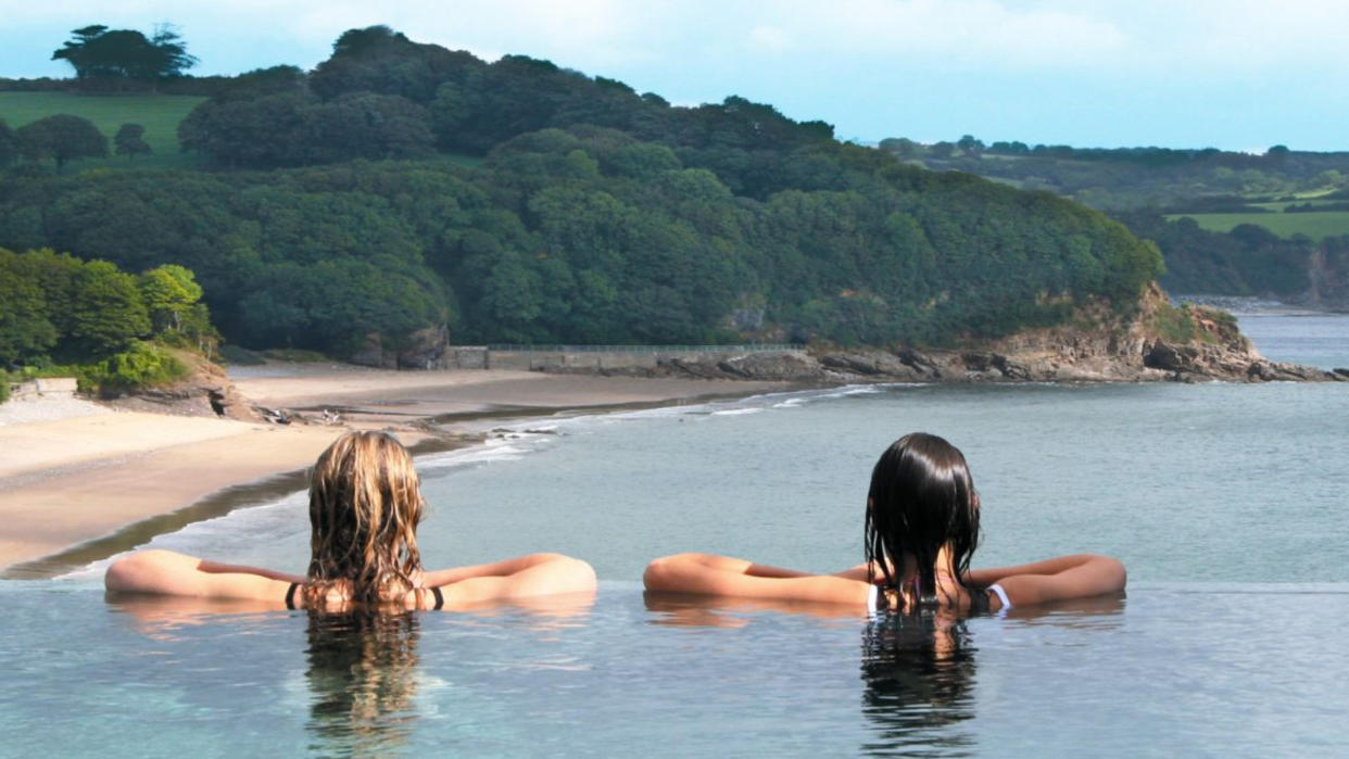  Rear view of two women looking from an infinity pool towards a bay and trees. 