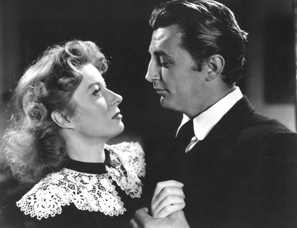 Greer Garson and Robert Mitchum stare into each other's eyes while holding hands, dressed in period attire with lace detail and formal suits