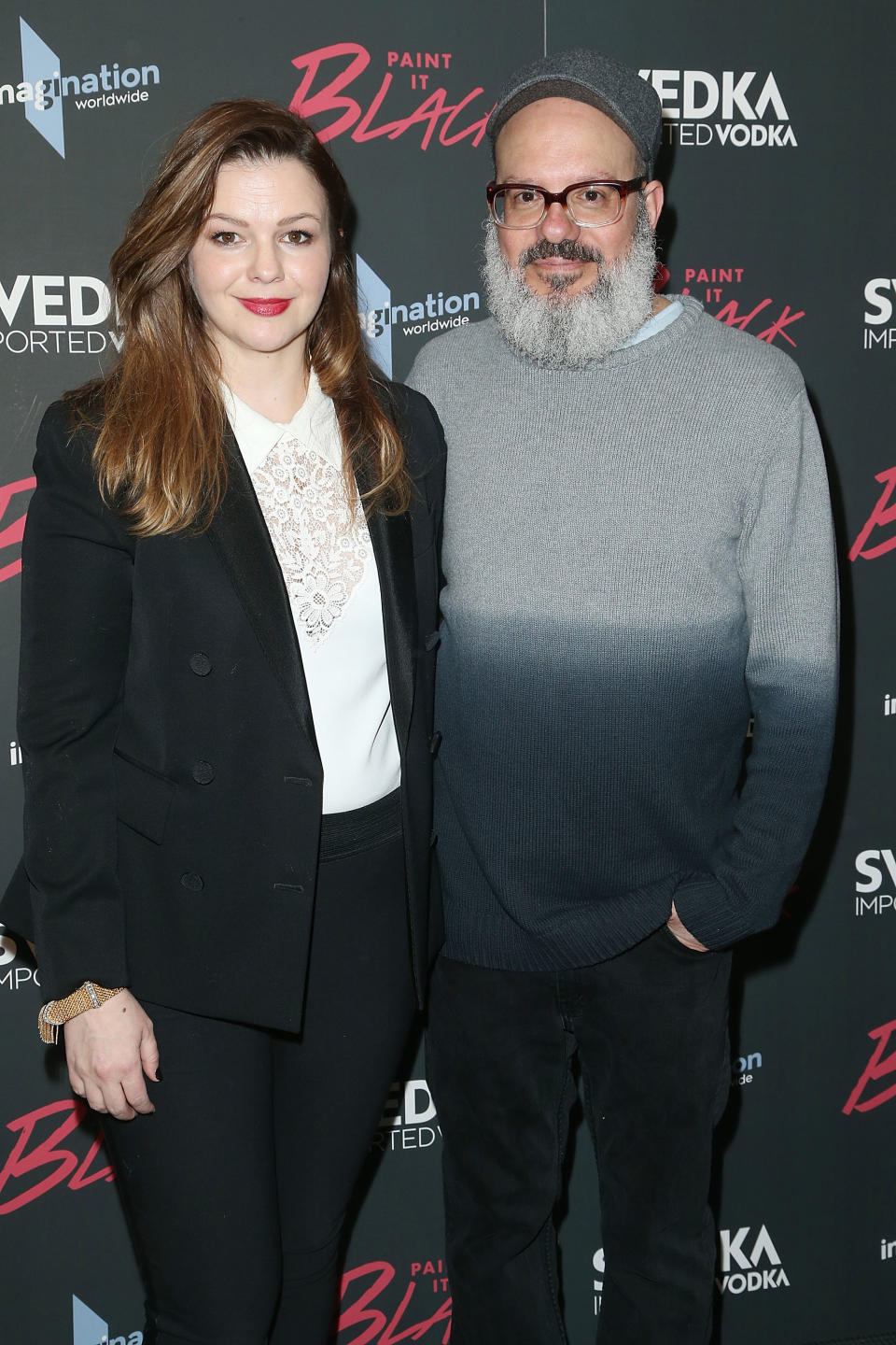 Two people posing together.  One is wearing a black blazer and white top, and the other is wearing a gray sweater.They participate in events with a brand background