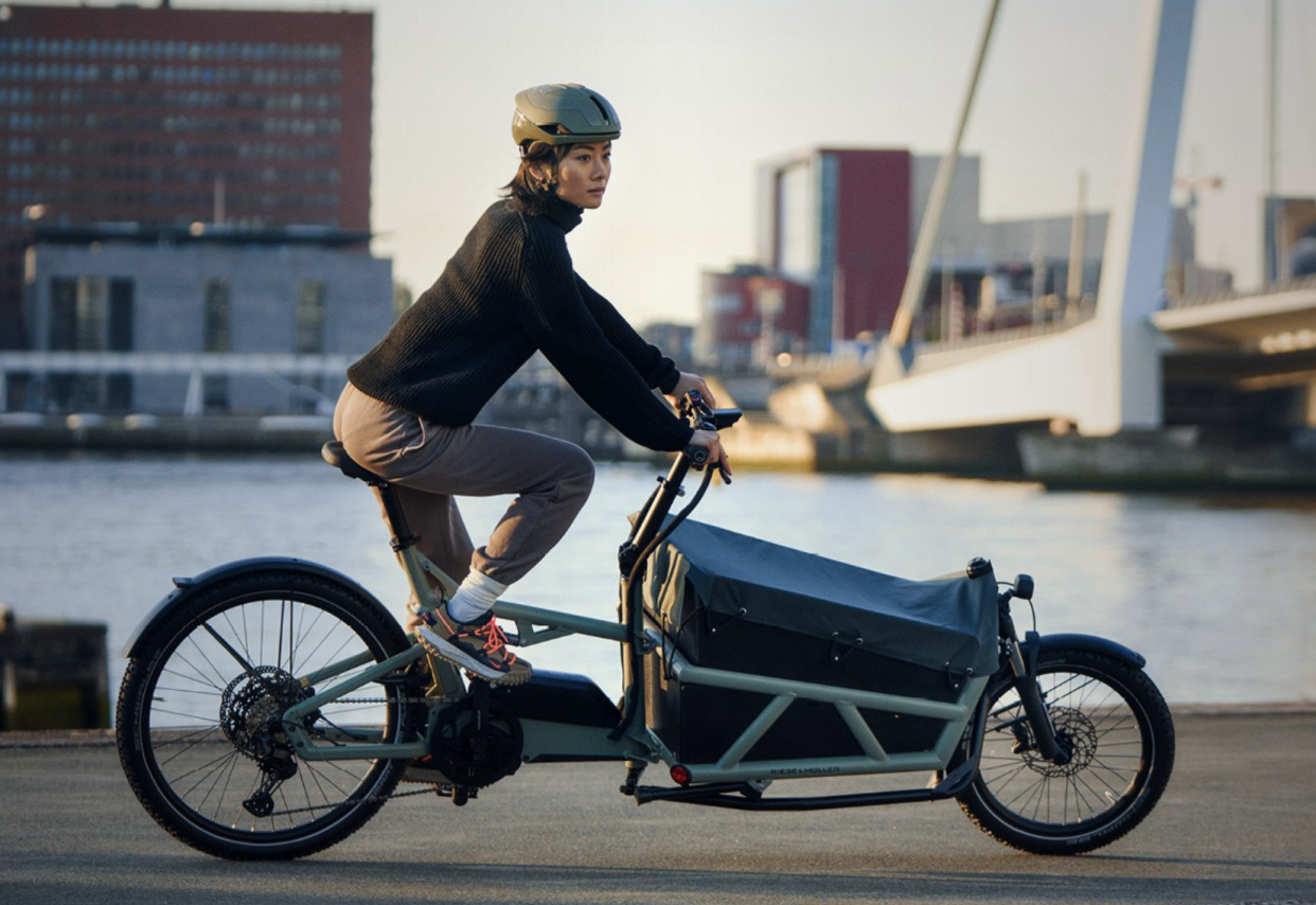  A woman dressed in black rides a front loading Riese and Muller cargo bike along a dock front. 