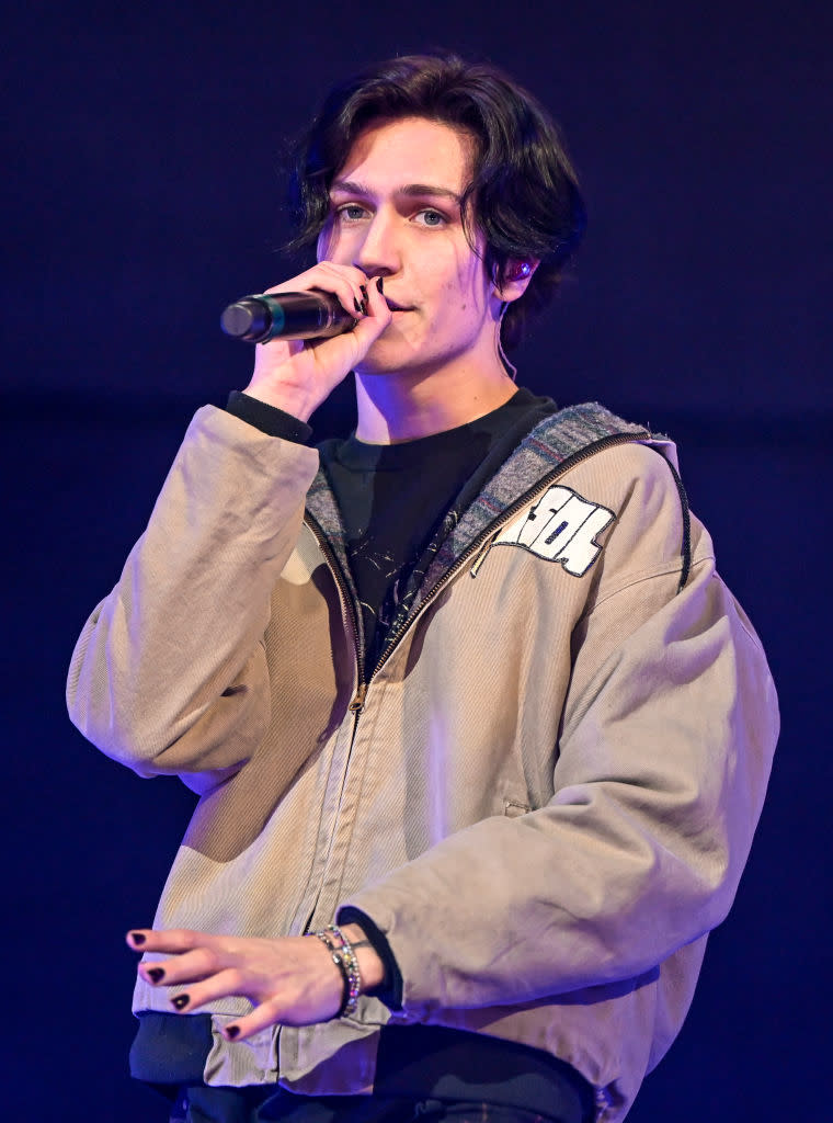 Huddy performing on stage with microphone, wearing a casual zip-up jacket