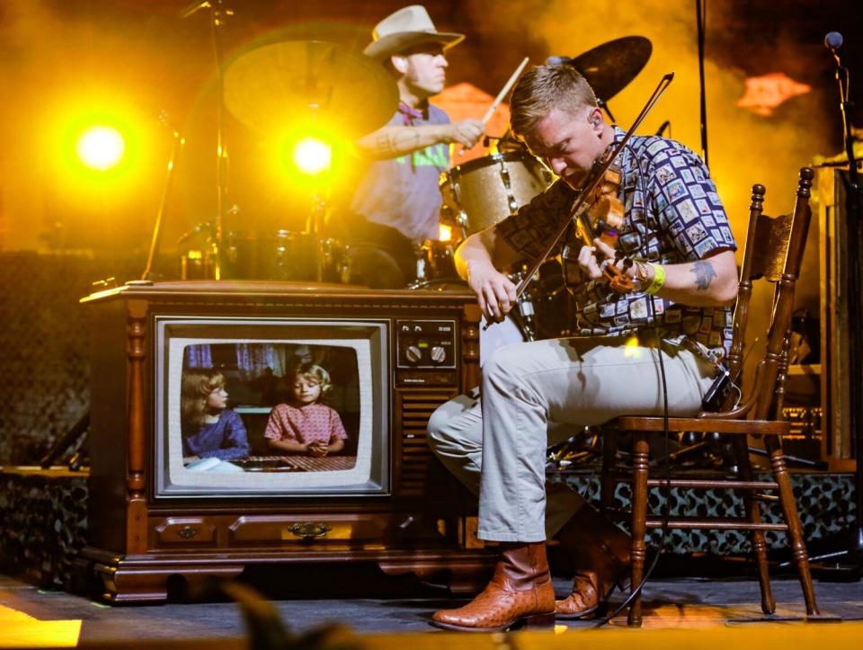 Two musicians onstage, one playing drums, the other seated on a chair next to an old-fashioned TV showing two young girls.