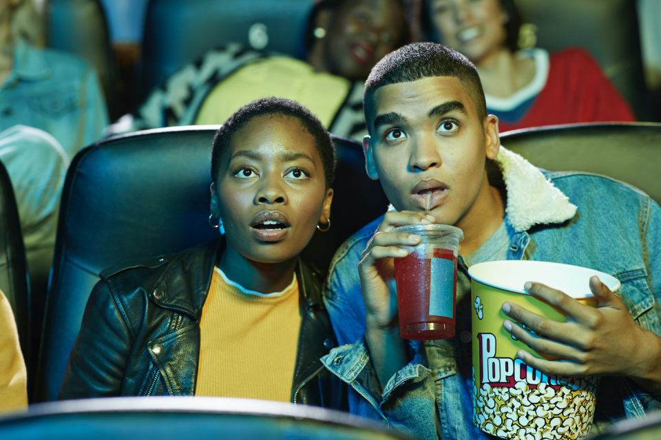 surprised young man drinking soda while watching movie with friend in cinema hall