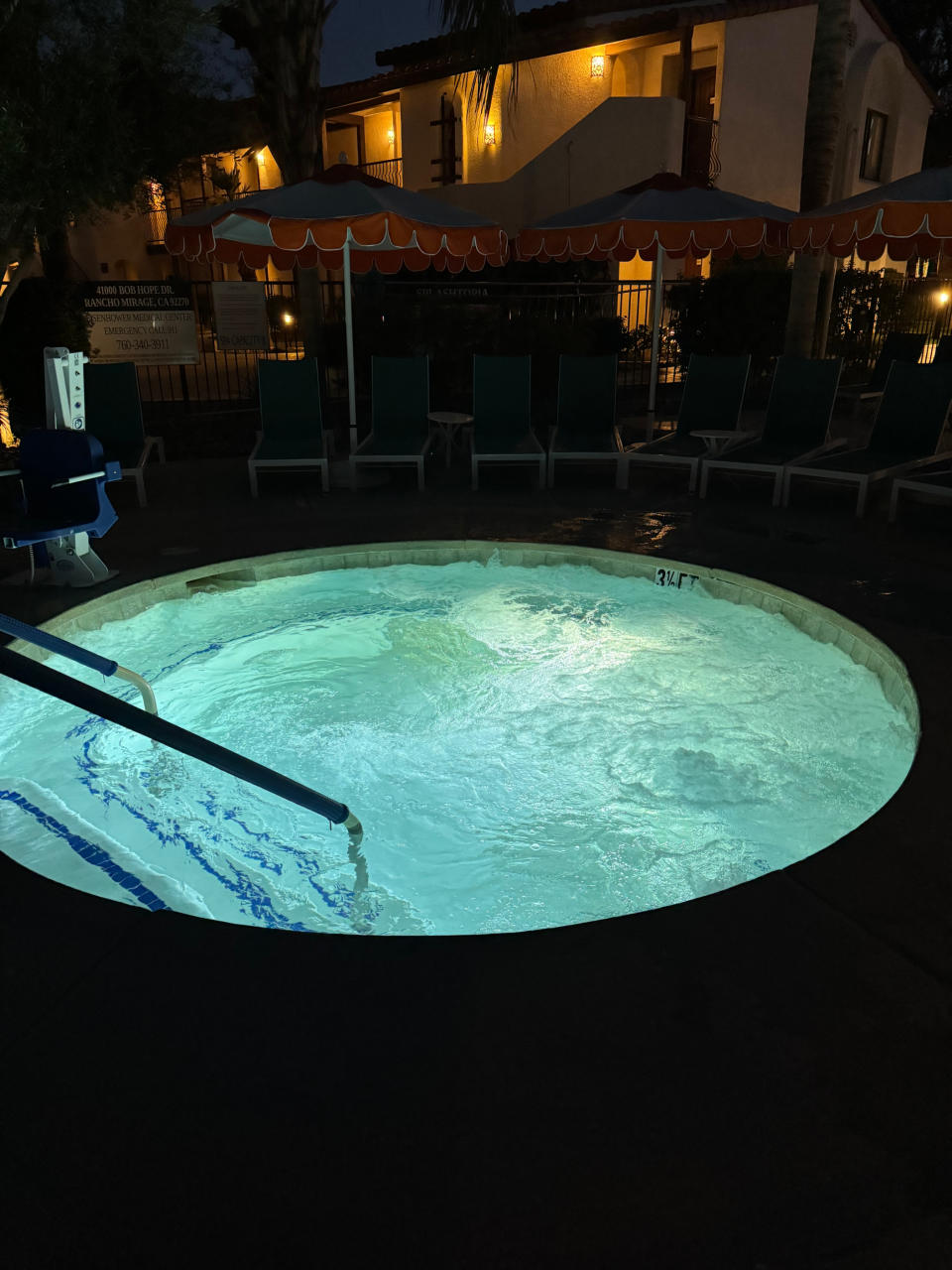 Nighttime setting of an outdoor hot tub with steam, surrounded by reclining chairs and pool umbrellas