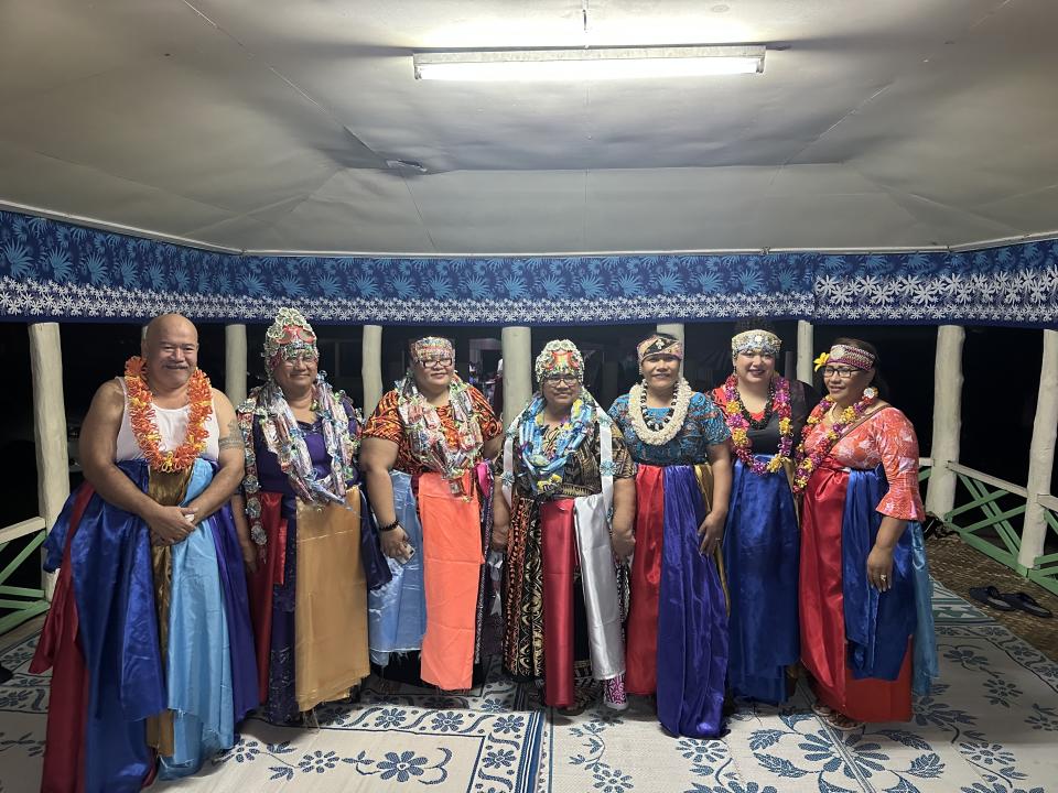 A group of seven individuals in traditional Polynesian attire, adorned with leis and headpieces, stand together in a decorated shelter