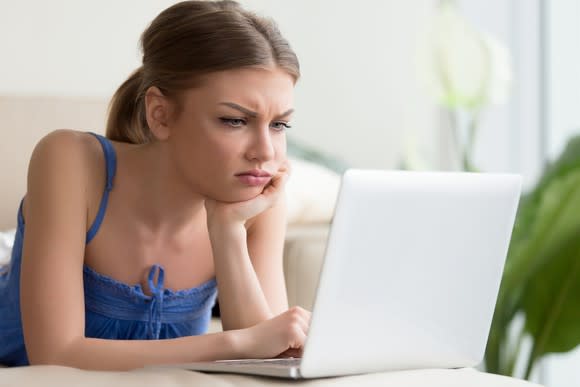 A woman rests her chin on her hand, appearing frustrated, as she looks at her laptop.