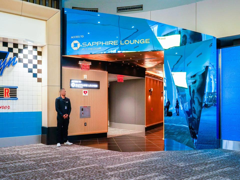 A man stands at the entrance of a blue-mirrored entrance to an airport lounge.