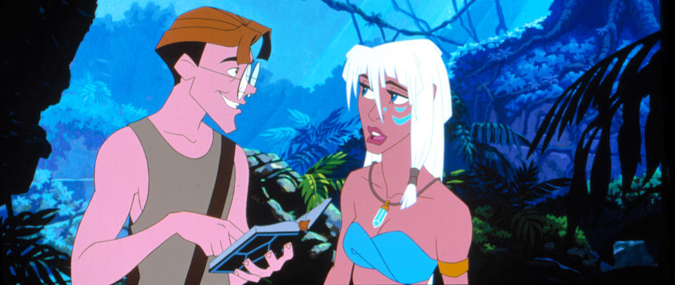 Milo Thatch, wearing a brown tank top and glasses, shows a book to Kida, who has white hair and is wearing a blue bandeau top and jewelry, in a lush jungle setting