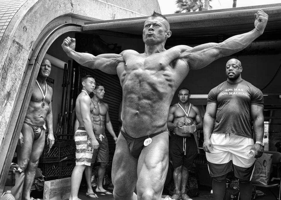 <p>A bodybuilder practices a pose backstage at the Mr. and Mrs. Muscle Beach Competition as a group of wary competitors watch him. Once the home gym of bodybuilders Arnold Schwarzenegger and Lou Ferrigno, the facility still serves the community as an iconic but accessible outdoor weight room. With a yearly membership cost of only $170 and aging gym equipment, the community gym has not yet succumbed to the gentrificationthat surrounds it. (© Dotan Saguy) </p>