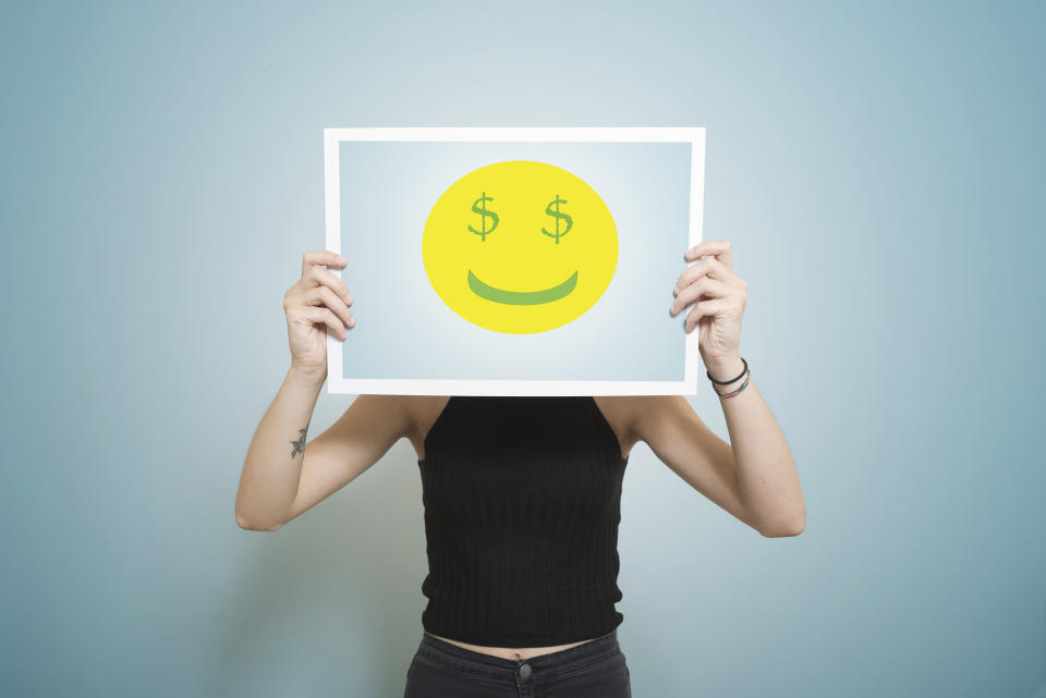 Woman holds up a poster of a smiley face with dollar signs as eyes over her face. She is wearing a black t-shirt and the background is blue.
